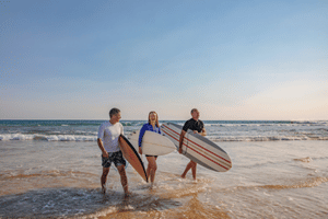 Three people walking out of the waves, chatting to each other and each holding a surfboard. Source: Destination Gold Coast